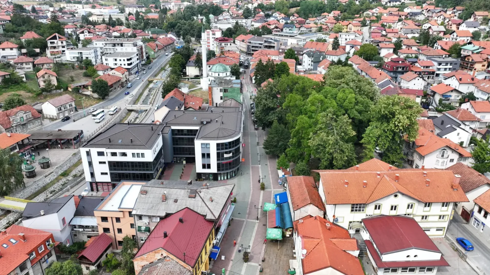 Panoramski pogled iz zraka na gradsku ulicu sa modernim staklenim zgradama, tradicionalnim kućama sa crvenim krovovima, drvećem i automobilima u pokretu.