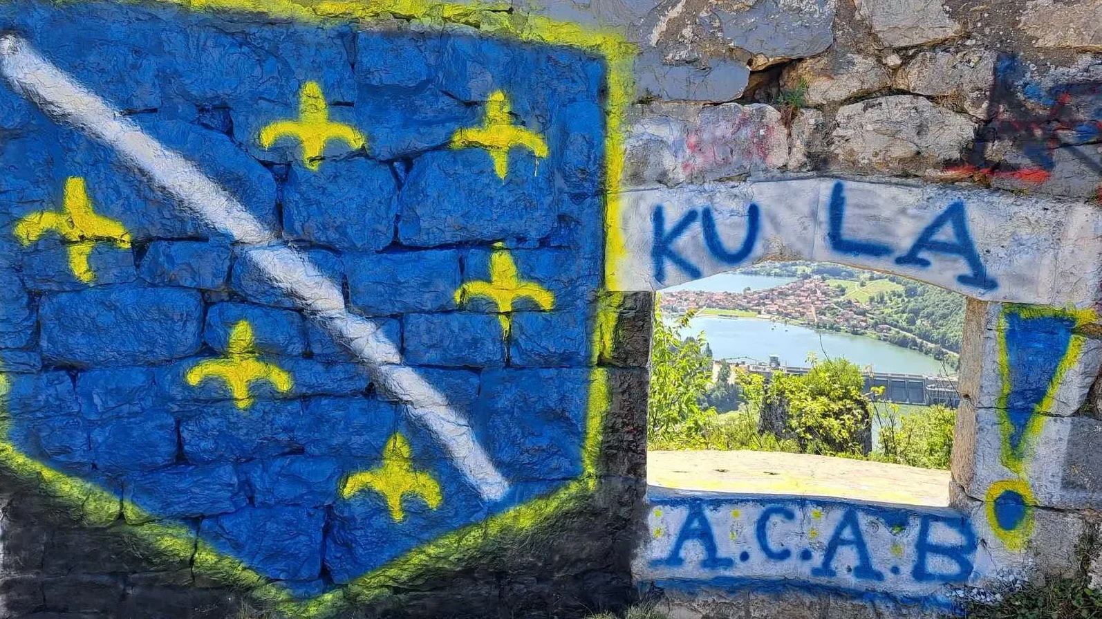 Grafiti sa ljiljanima i kosom crtom na plavoj podlozi naslikani na kameni zid, te natpisi "KULA" i "A.C.A.B." iznad i ispod otvora kroz koji se vidi rijeka i naselje.