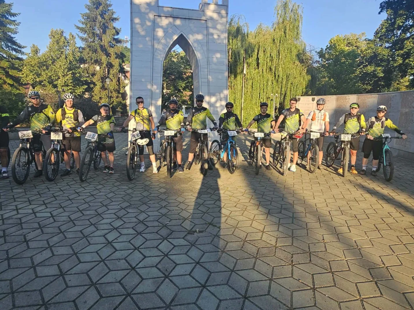 Grupa biciklista u sportskim majicama i kacigama pozira sa svojim biciklima ispred bijelog spomenika.