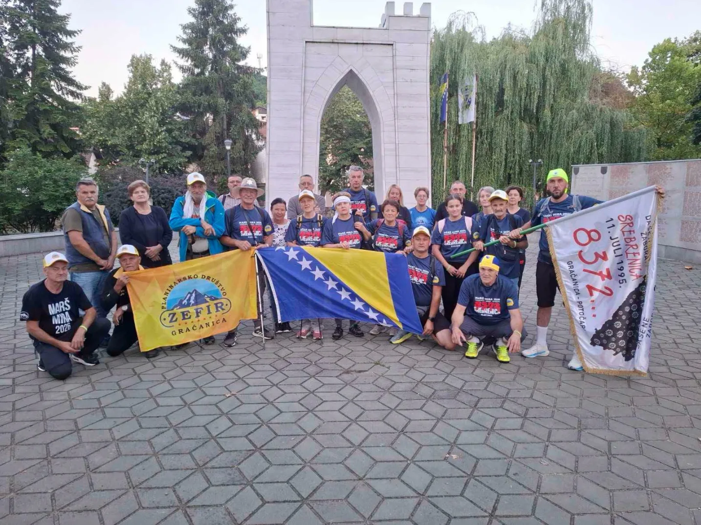Grupa ljudi, odraslih i djece, stoji ispred bijelog spomenika sa lukovima i drveta. Neki od njih drže zastave Bosne i Hercegovine i zastavu s natpisom "Srebrenica 11. juli 1995. Gračanica - Potočari" i brojem 8372. Većina nosi majice s istim logom i natpisima "MARŠ MIRA 2026" ili slično. Pozadina je djelimično zaklonjena drvećem i spomenikom.