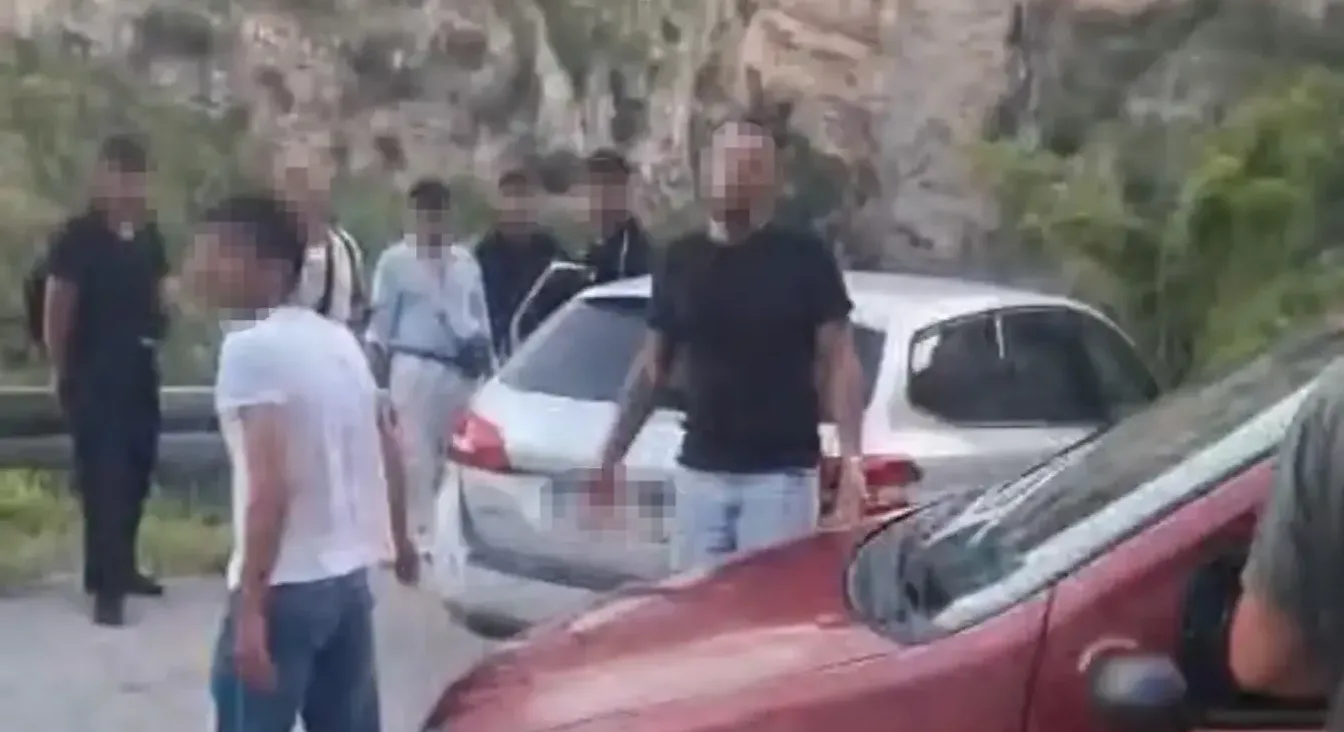 Grupa ljudi okupila se pored parkiranih automobila naizgled uz rub puta, s brdovitom pozadinom obraslom rastinjem.