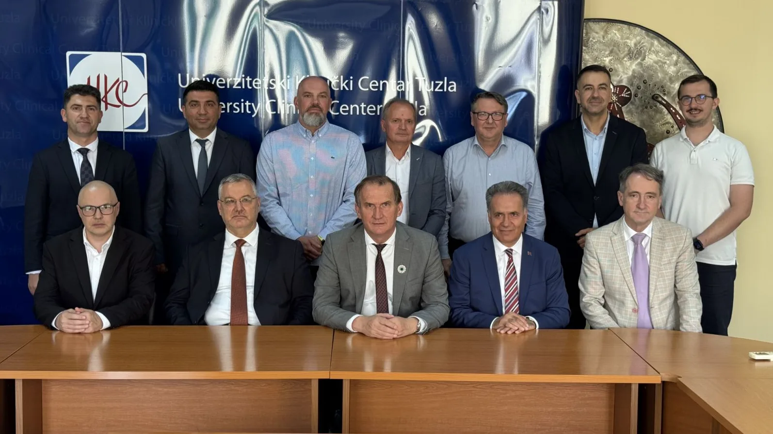 Grupa muškaraca u odijelima i košuljama pozira za fotografiju ispred plavog panoa s logom Univerzitetskog kliničkog centra Tuzla.