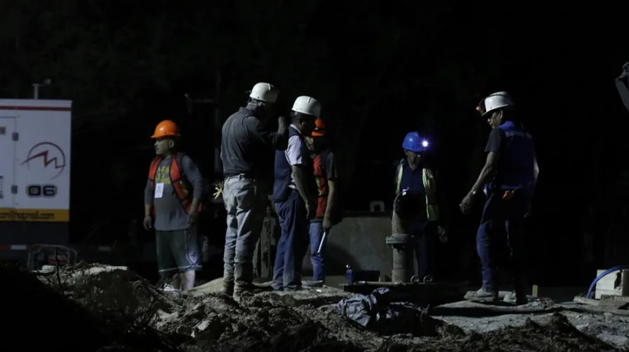 Grupa radnika u zaštitnim odijelima i kacigama na gradilištu noću, osvijetljeni reflektorima i lampama na kacigama.