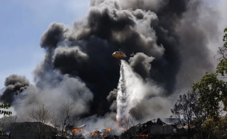 Helikopter sa žutom i plavom bojom ispušta vodeni mlaz iznad gustog crnog dima koji suklja iz zapaljenih objekata prizemlje.