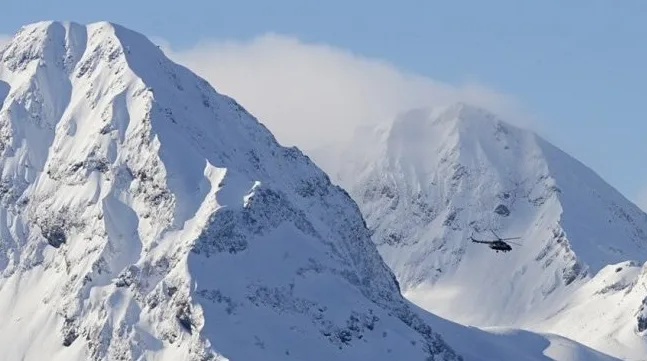 Helikopter leti iznad snijegom prekrivenih planina pod plavim nebom.