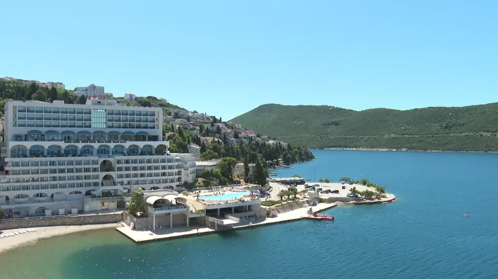 Veliki bijeli hotel sa terasama i balkonima smješten na obali mora, uz plavo more i zelene planine u pozadini. Ispred hotela nalazi se bazen sa ležaljkama i suncobranima.