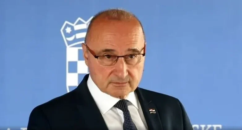 Portret muškarca u tamnom odijelu s bijelom košuljom i tamnom kravatom, koji nosi naočale. U pozadini se vidi dio grba Republike Hrvatske.