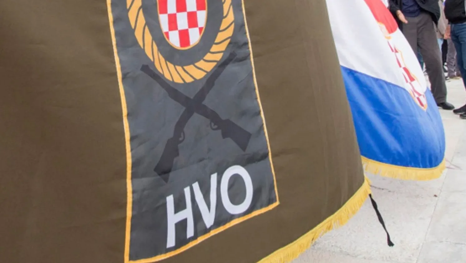 Zastava sa grbom Hrvatskog vijeća odbrane (HVO) i ukrštenim puškama, pored nje se vidi dio zastave Republike Hrvatske.