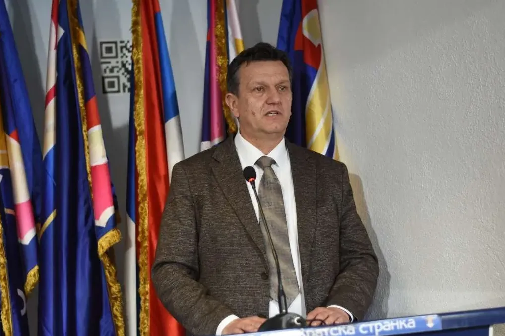 Predsjednik Hrvatske stranke, obučen u smeđe odijelo i bijelu košulju s kravatom, stoji za govornicom ispred niza zastava, uključujući zastave Bosne i Hercegovine, Republike Hrvatske i Evropske unije.