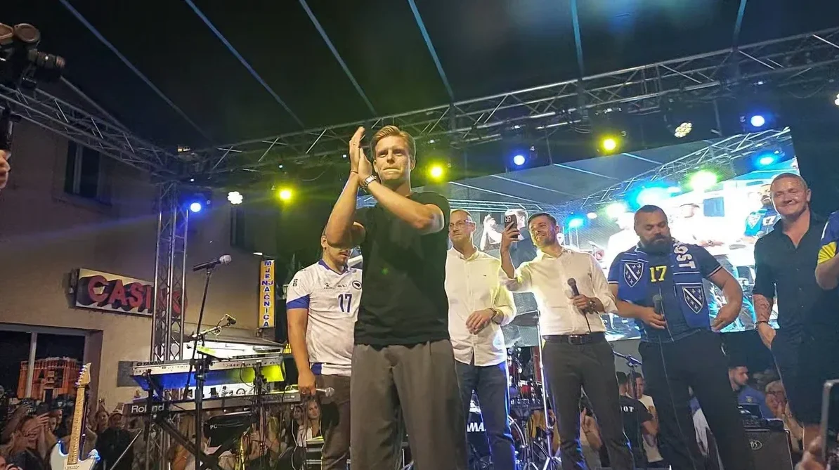 Čovjek na bini pljeska rukama, okružen drugim muškarcima u sportskoj odjeći i prisutnom publikom na koncertu na otvorenom.