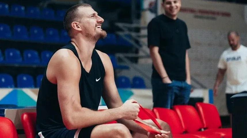 Nasmijani košarkaš u crnoj majici bez rukava sjedi na klupi i drži crvenu traku.