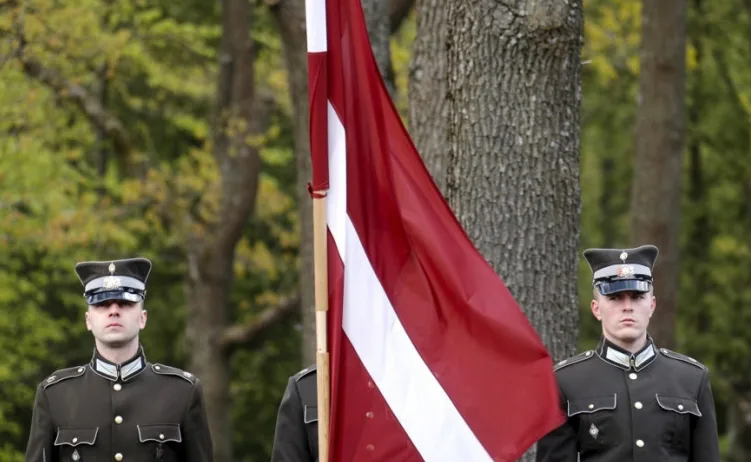 Dvije vojne osobe u uniformama stoje u počasnoj straži pored zastave Latvije.