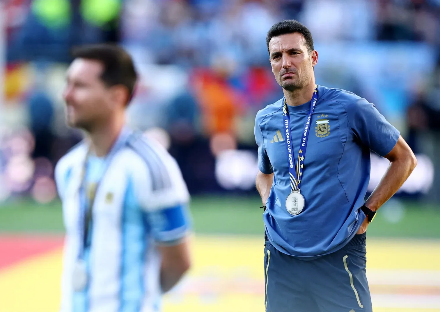 Lionel Scaloni, selektor argentinske fudbalske reprezentacije, nosi medalju i gleda prema terenu, dok je Lionel Messi u prvom planu, također s medaljom oko vrata.