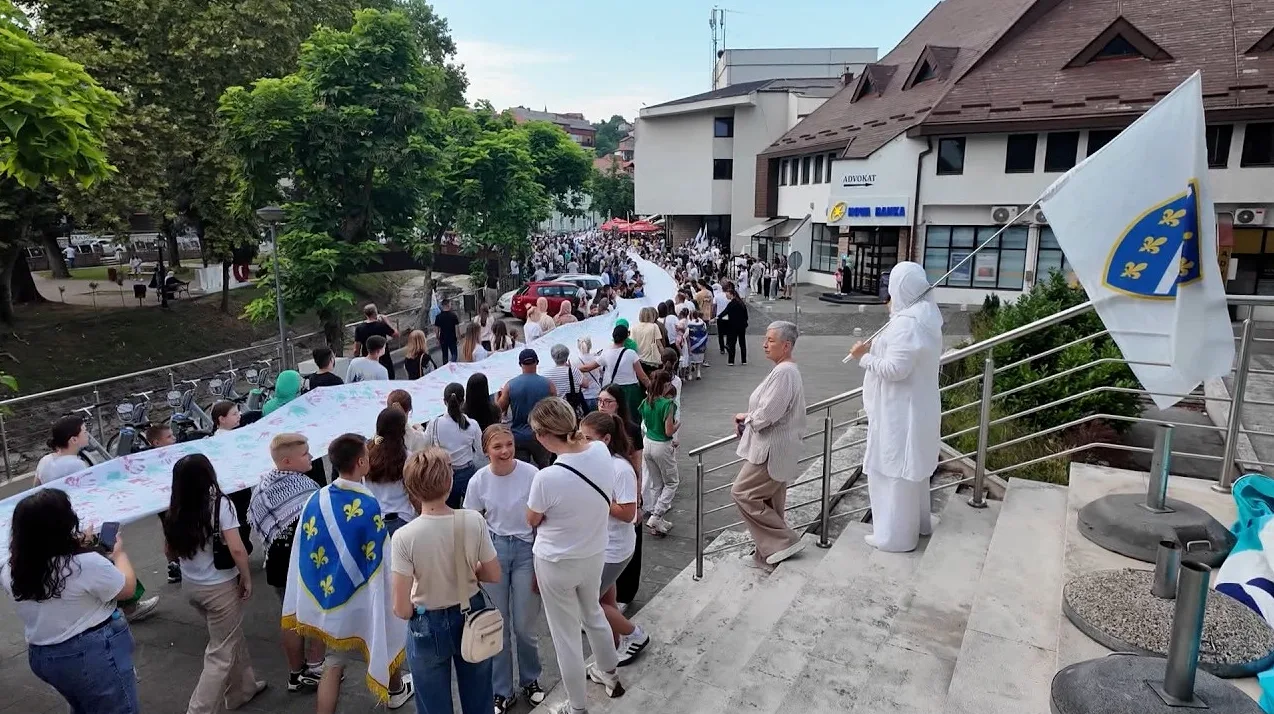 Grupa ljudi nosi dugačak bijeli transparent sa simbolima i natpisima, dok jedan učesnik nosi zastavu Bosne i Hercegovine sa ljiljanima.