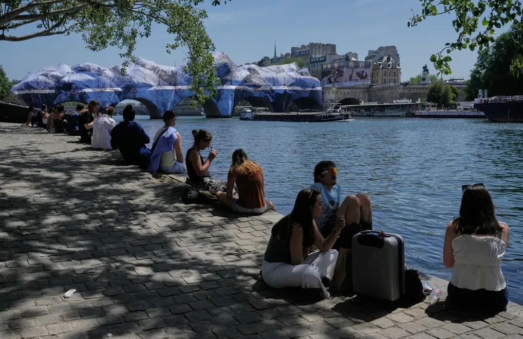 Grupa ljudi sjedi na obali rijeke Seine u Parizu, sa umjetničkom instalacijom u pozadini.