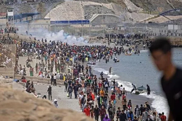 Grupa migranata na plaži u blizini granice, sa suzavcem u pozadini.