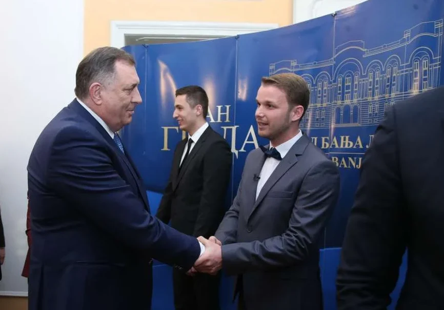 Predsjednik entiteta RS Milorad Dodik rukuje se sa mladim muškarcem u sivom odijelu s leptir mašnom, dok se iza njih vidi plava pozadina sa bijelim natpisom i ilustracijom zgrade.