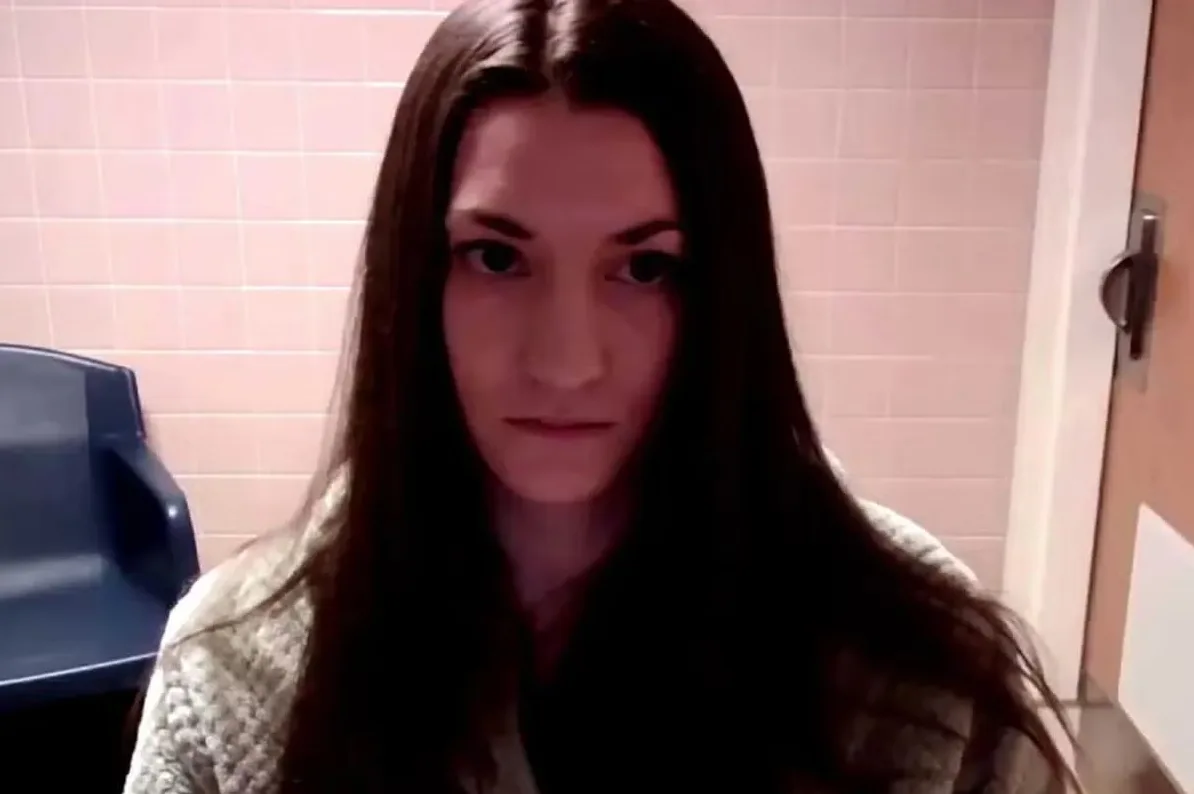 Portret mlade žene sa dugom, tamnom kosom koja pada preko ramena, gleda direktno u kameru blagim izrazom lica. Pozadina je obložena svjetlo roze ili boje breskve, a desno od nje vidi se dio vrata i kvaka.