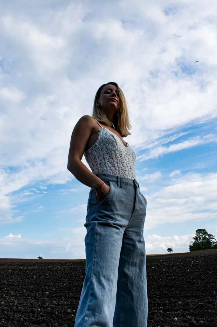 Mlada žena sa plavom kosom, obučena u bijeli top i farmerke, stoji pod vedrim nebom i gleda u daljinu, u polju sa tamnom zemljom.