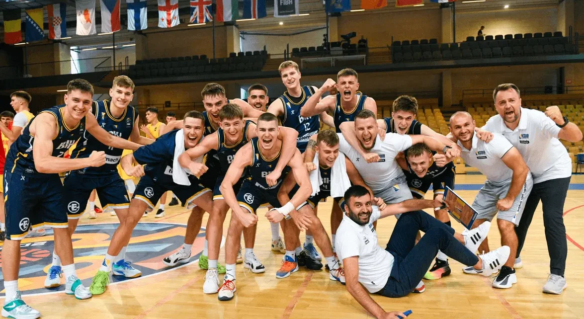 Grupa mladih košarkaša Bosne i Hercegovine, u tamnoplavim dresovima, drži pehar i slavi pobjedu na parketu sportske dvorane.