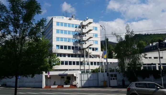 Fotografija prikazuje modernu bijelu zgradu sa redovima plavih prozora, karakterističnim stepeništem na vanjskoj strani zgrade i zastavom Bosne i Hercegovine istaknutom pored ulaza.