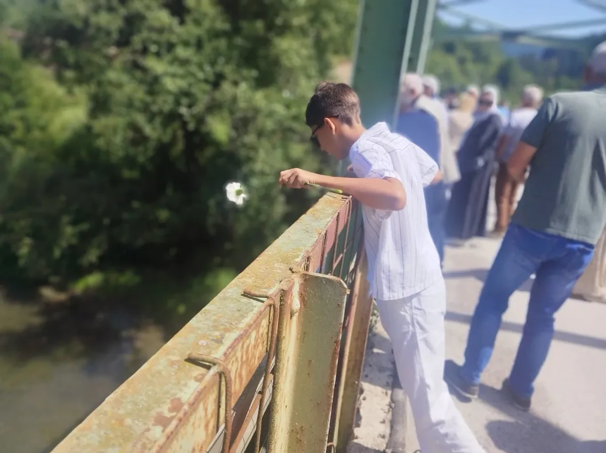 Mladić u bijeloj košulji i bijelim hlačama s naočalama stoji na mostu i baca bijeli cvijet prema rijeci.