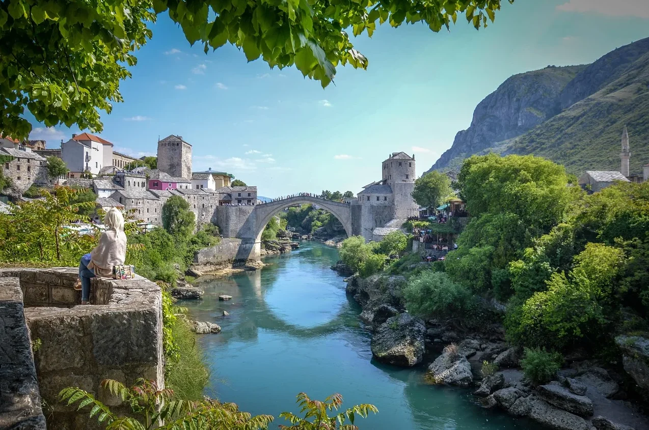Pogled na Stari most u Mostaru iznad smaragdne rijeke Neretve, sa starim kamenim kućama i zelenilom koje okružuje obalu.