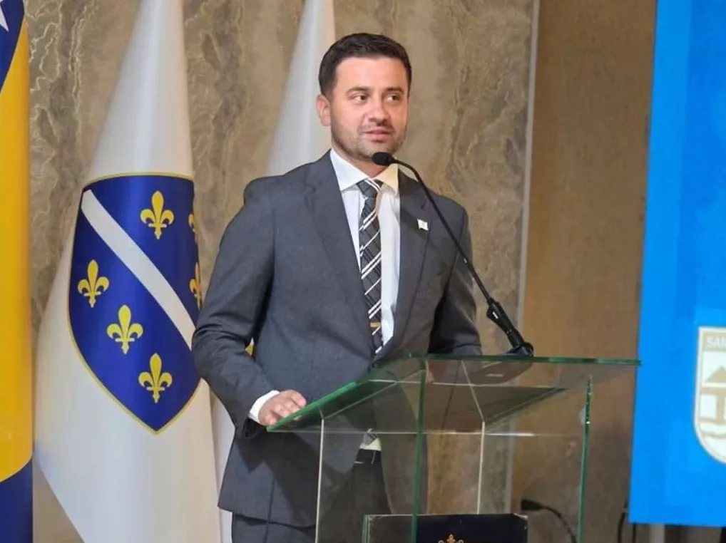 Političar ili javna ličnost, obučen u tamni odijelo i bijelu košulju sa prugastom kravatom, stoji iza prozirnog podijuma i govori u mikrofon. U pozadini se vide zastave Bosne i Hercegovine i još jedna sa grbom.