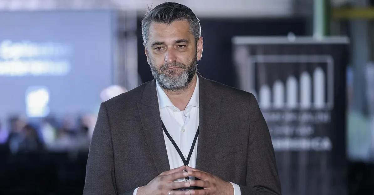 Portret muškarca srednjih godina sa sijedom bradom i kosom, obučenog u tamni sako i bijelu košulju, kako stoji ispred bine na konferenciji.