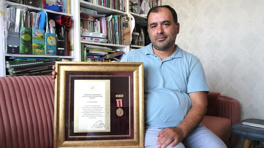 Muškarac u plavoj majici sjedi i drži uokvirenu diplomu i odlikovanje.