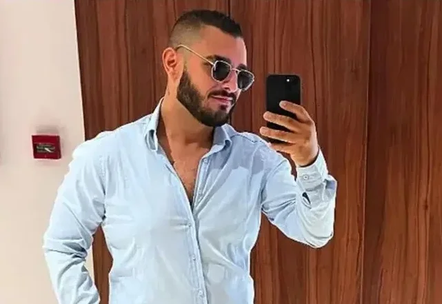 Mlad muškarac sa urednom bradom i naočarima za sunce slika se telefonom, obučen u svijetloplavu košulju.