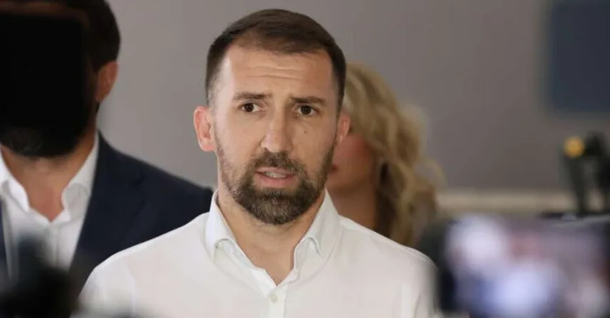 Portret muškarca sa kratkom smeđom kosom i bradom, obučenog u bijelu košulju, dok se u pozadini naziru druge osobe i oprema za snimanje.