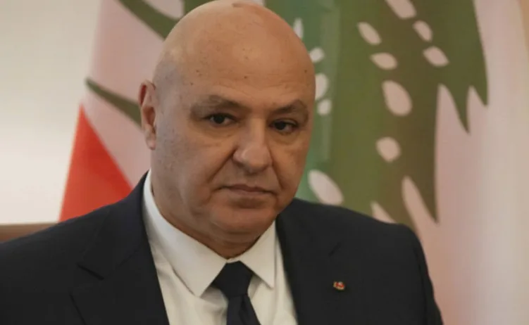 Muskarac u tamnom odijelu i bijeloj kosulji sa tamnom kravatom, kratko osisane glave, gleda u kameru. U pozadini se vidi dio zastave.