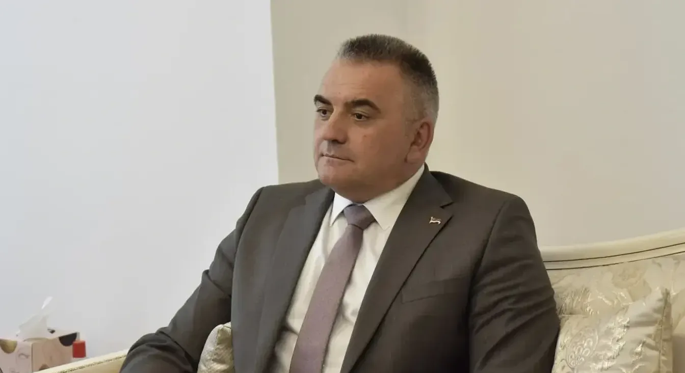 Muškarac u tamnom odijelu i ljubičastoj kravati sjedi i gleda u stranu.