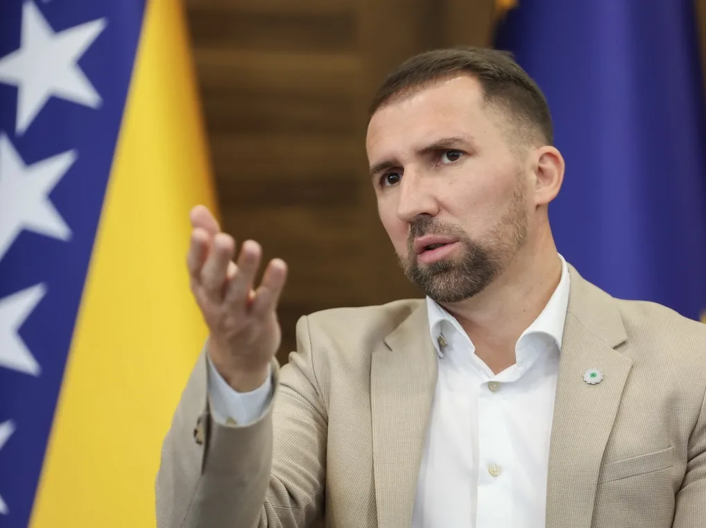 Muškarac u bež sakou i bijeloj košulji govori sa podignutom rukom, ispred zastave Bosne i Hercegovine.