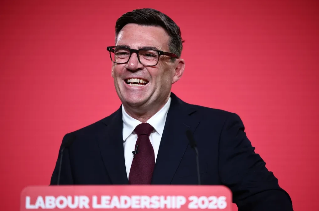Čovjek u tamnom odijelu, bijeloj košulji i tamnoj kravati, nosi crne naočale i smije se dok stoji iza govornice s natpisom 'Labour Leadership 2026'.