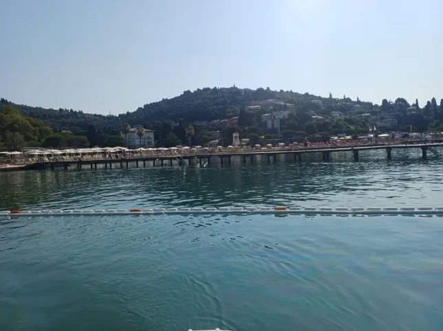 Pogled na morsku obalu sa dugačkim drvenim šetalištem prepunim ležaljki i suncobrana, dok se u pozadini uzdižu zelena brda sa gradnjom.