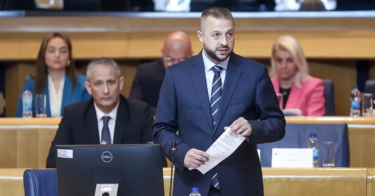 Političar u tamnoplavom odijelu i sa prugastom kravatom drži papire i govori u sali za sastanke.