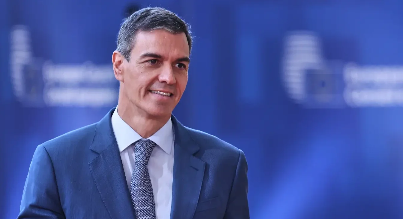 Portret Pedra Sancheza u plavom odijelu s bijelom košuljom i plavom kravatom sa sitnim uzorkom, gleda udesno s blagim osmijehom. Pozadina je tamnoplave boje sa zamućenim bijelim svjetlosnim efektima.
