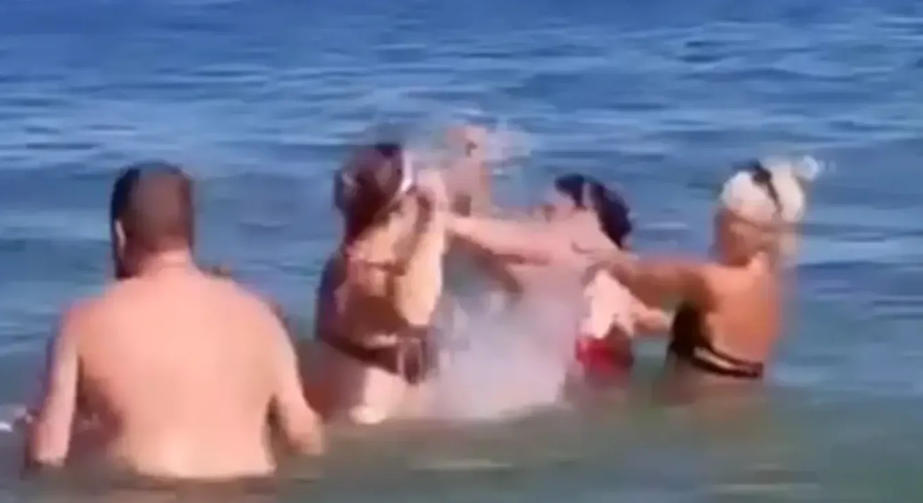 Dvije žene, plavuša i brineta, svađaju se u plićaku mora, dok ih sa strane promatraju dva muškarca.