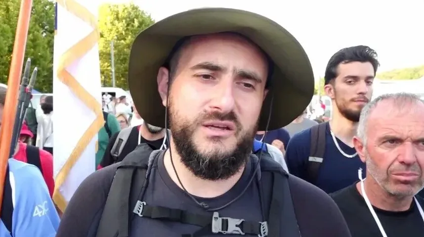 Muškarac sa šeširom i bradom gleda prema kameri, okružen drugim ljudima i zastavama.