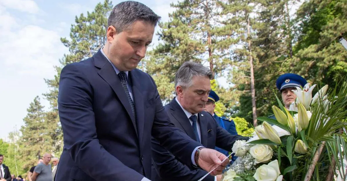 Dva muškarca u odijelima polažu vijenac bijelog cvijeća ispred drveća.