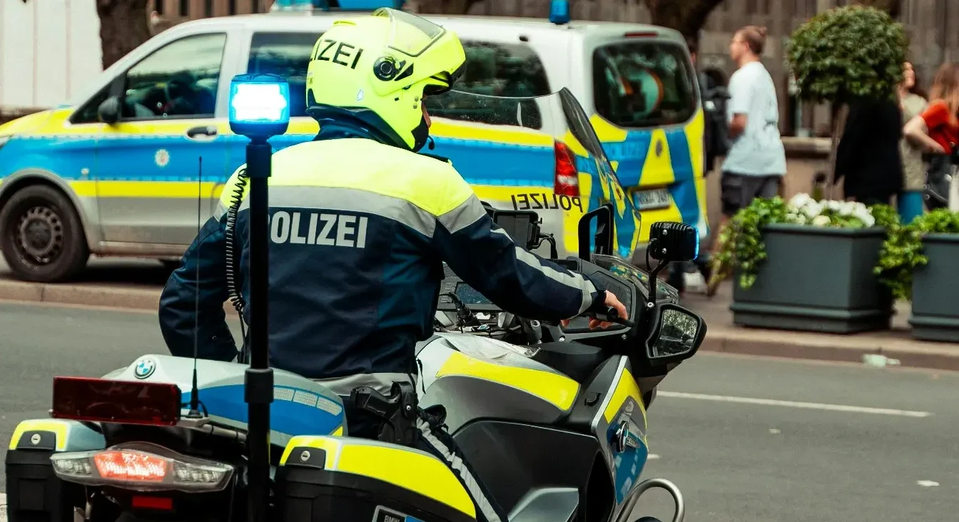Policajac u punoj opremi, na motociklu sa plavim rotacionim svjetlom, ispred policijskog automobila.
