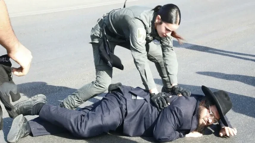 Policajka u zelenoj uniformi savladava muškarca u crnom odijelu i šeširu, koji leži na asfaltu.