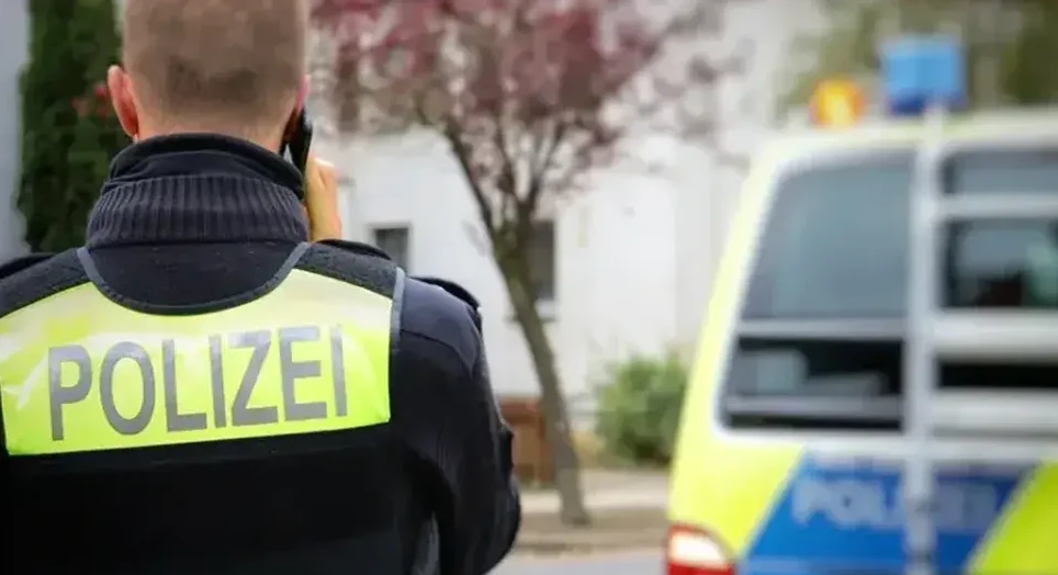 Policajac u uniformi s natpisom 'Polizei' na prsluku razgovara telefonom ispred policijskog vozila.