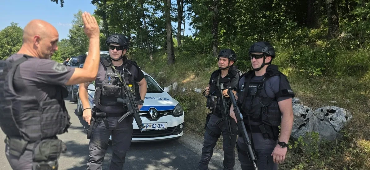 Pripadnici specijalne policije u punoj opremi sa automatskim puškama stoje pored policijskog automobila u šumovitom predjelu.