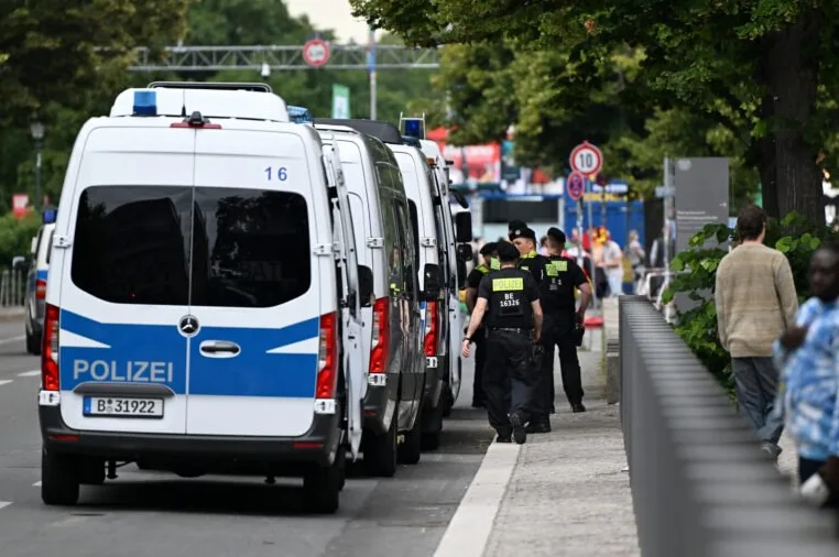 Red policijskih kombija s upaljenim rotacijskim svjetlima i policijski službenici u uniformama na trotoaru.