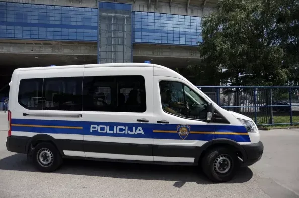 Bijelo policijsko kombi vozilo sa plavim i žutim prugama, sa natpisom "POLICIJA" i grbom, parkirano je ispred modernog objekta sa reflektirajućim plavim prozorima.