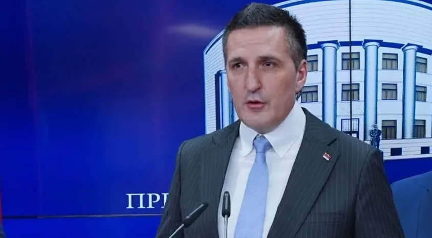 Portret političara u tamnom odijelu sa prugama i plavom kravatom kako govori pred mikrofonom.