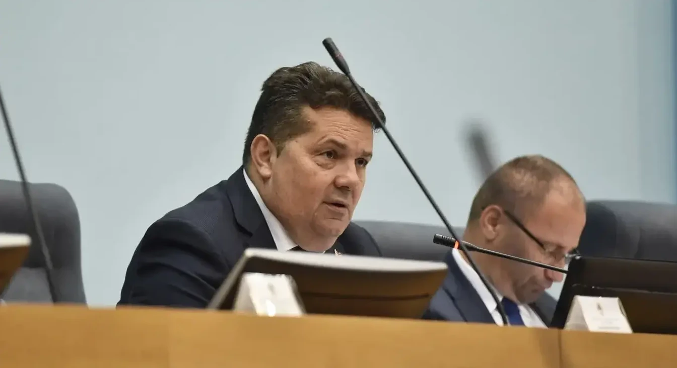 Političar u tamnom odijelu govori za govornicom sa mikrofonom ispred sebe.